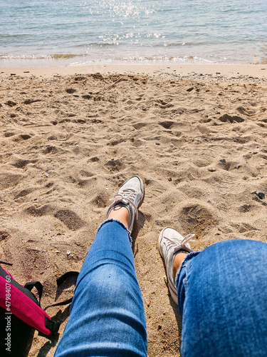 Woman legs in white sneakers and jeans on yellow sand beach in summer day. A woman is sitting on the seashore. Rest at the sea. Ocean clear water seafoam, yellow sand, small wawes. Summer background.