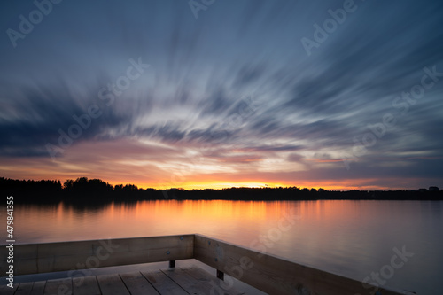 Fototapeta Naklejka Na Ścianę i Meble -  Panoramic Tranquil golden hour cloud above the forest lake at sunset. Dramatic cloudscape. Long exposure on the water