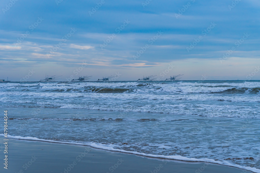 Scena con il mare con le onde e la spiaggia e dei padelloni sullo sfondo a Marina di Ravenna, in Italia, in inverno. Viaggiare. Vacanze. Destinazione. Paesaggio marino.