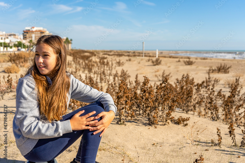Pretty pre-adolescent Hispanic girl sitting on top of her soccer ball in the sand of Burriana beach bathed by the waters of the Mediterranean.