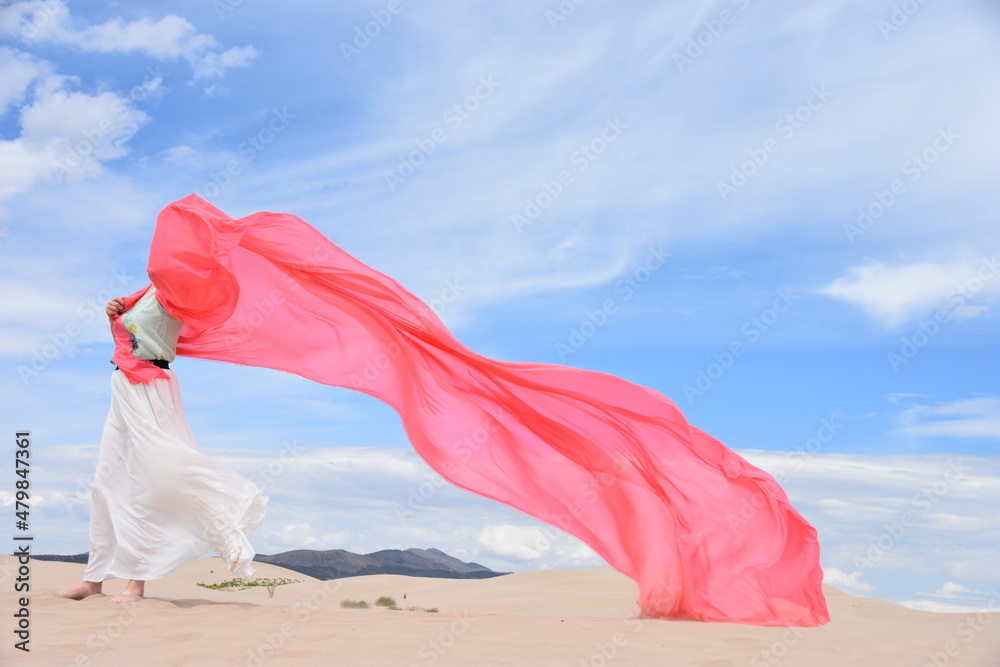 Young woman standing in sand dunes with a wide and large coral pink color textile silk scarf around her on a windy day. Young woman standing in sand dunes with a wide and large coral pink color textile silk scarf around her on a windy day.