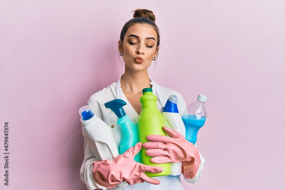 Beautiful brunette young woman wearing cleaner apron holding cleaning products looking at the camera blowing a kiss being lovely and sexy. love expression.