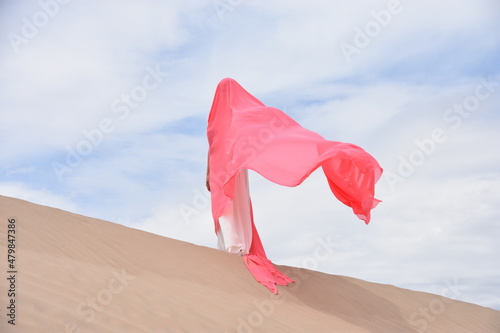 Young woman standing in sand dunes with a wide and large coral pink color textile silk scarf around her on a windy day.