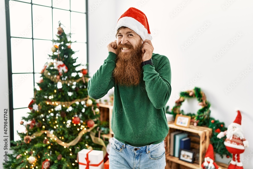 Redhead man with long beard wearing christmas hat by christmas tree covering ears with fingers with annoyed expression for the noise of loud music. deaf concept. Redhead man with long beard wearing christmas hat by christmas tree covering ears with fingers with annoyed expression for the noise of loud music. deaf concept.
