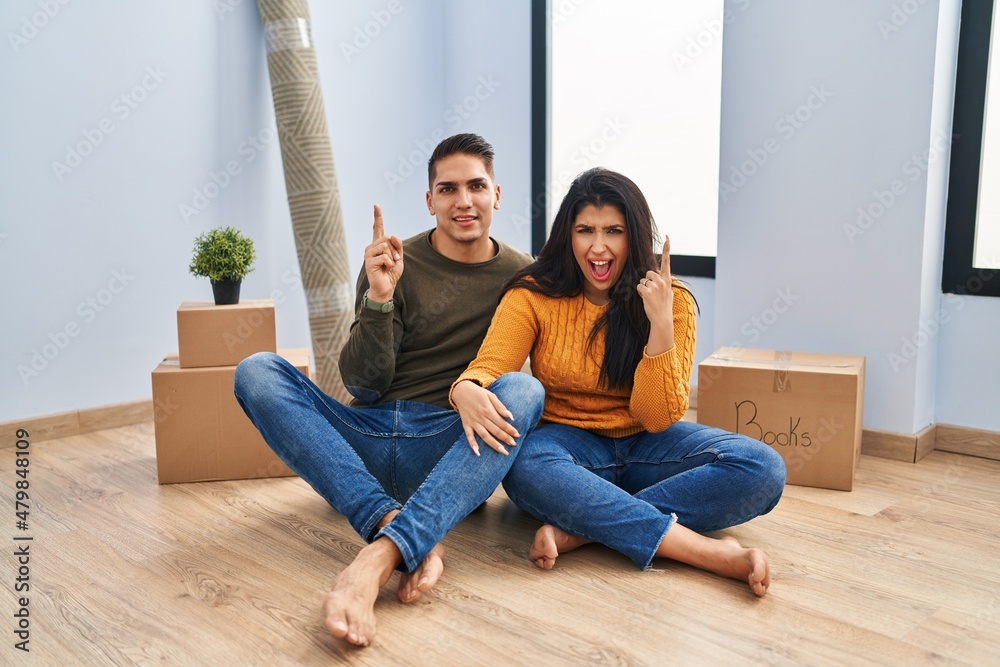Young couple sitting on the floor at new home pointing finger up with successful idea. exited and happy. number one. Young couple sitting on the floor at new home pointing finger up with successful idea. exited and happy. number one.