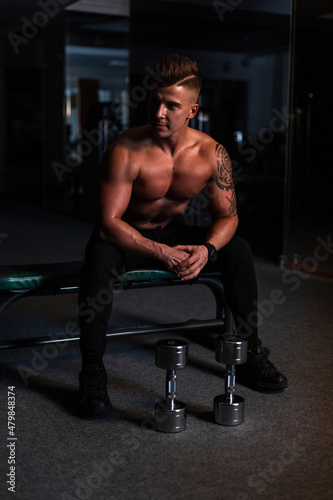 Handsome young sporty man with naked body and muscles sits near dumbbell in the gym in the dark. Workout