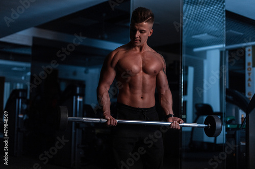 Handsome strong young sports man with a haircut and a naked torso with muscles doing a workout in the hall on a dark background. Male sexy body