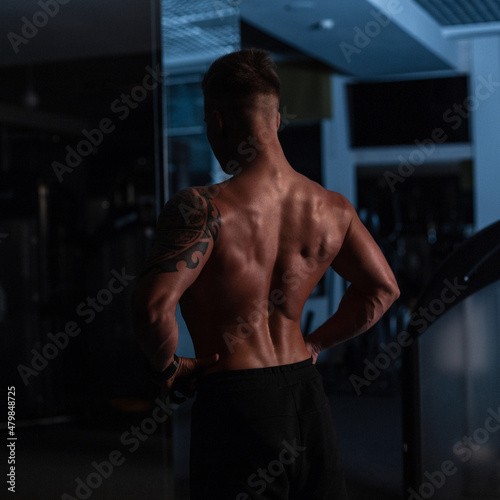 young handsome sports guy with a muscular naked torso stands with his back to the camera in the sport gym