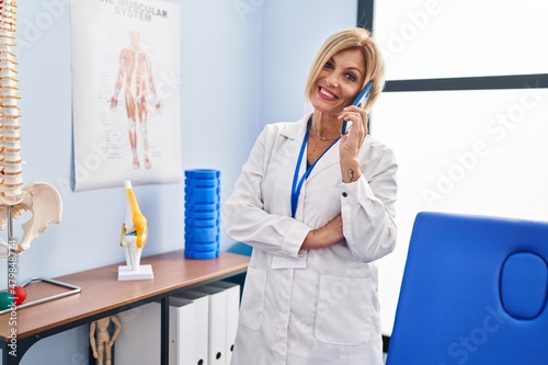 Middle age blonde woman wearing physiotherapist uniform talking on the smartphone at physiotherapy clinic
