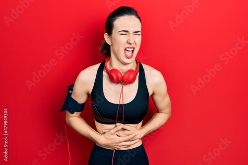 Young hispanic girl wearing gym clothes and using headphones with hand on stomach because nausea, painful disease feeling unwell. ache concept.