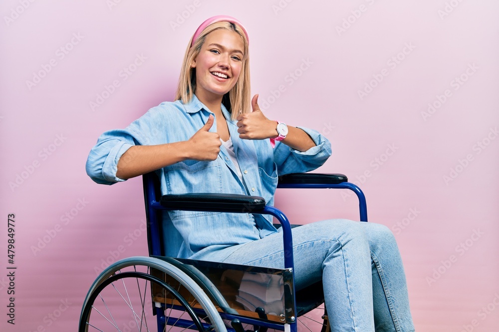 Beautiful blonde woman sitting on wheelchair success sign doing positive gesture with hand, thumbs up smiling and happy. cheerful expression and winner gesture. Beautiful blonde woman sitting on wheelchair success sign doing positive gesture with hand, thumbs up smiling and happy. cheerful expression and winner gesture.