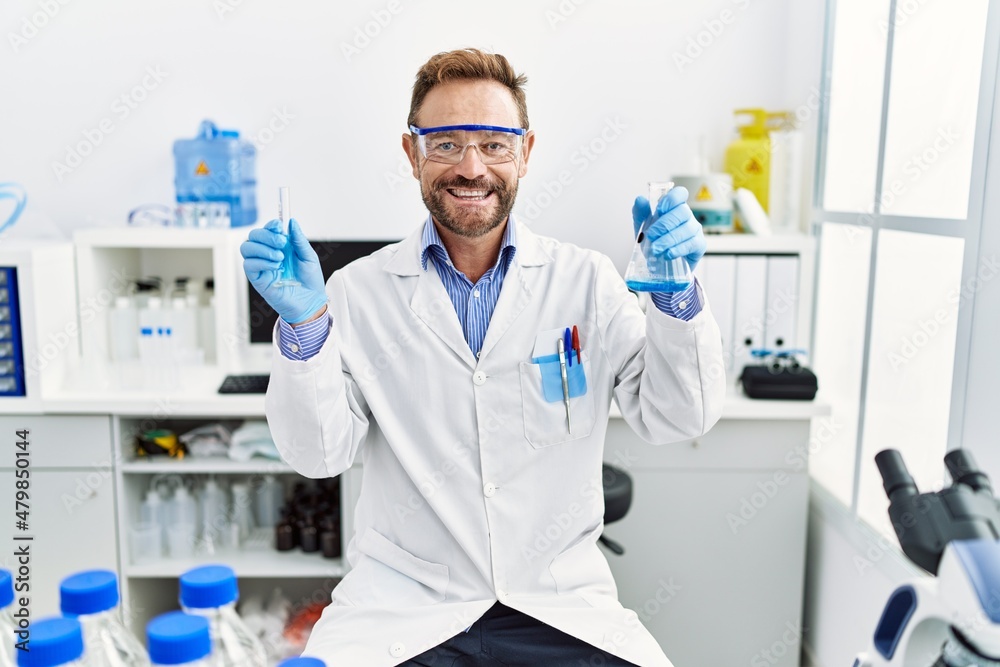 Middle age man working at scientist laboratory holding chemical products smiling with a happy and cool smile on face. showing teeth. Middle age man working at scientist laboratory holding chemical products smiling with a happy and cool smile on face. showing teeth.