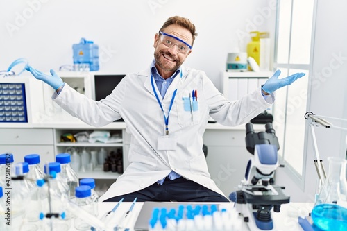 Middle age man working at scientist laboratory clueless and confused expression with arms and hands raised. doubt concept.