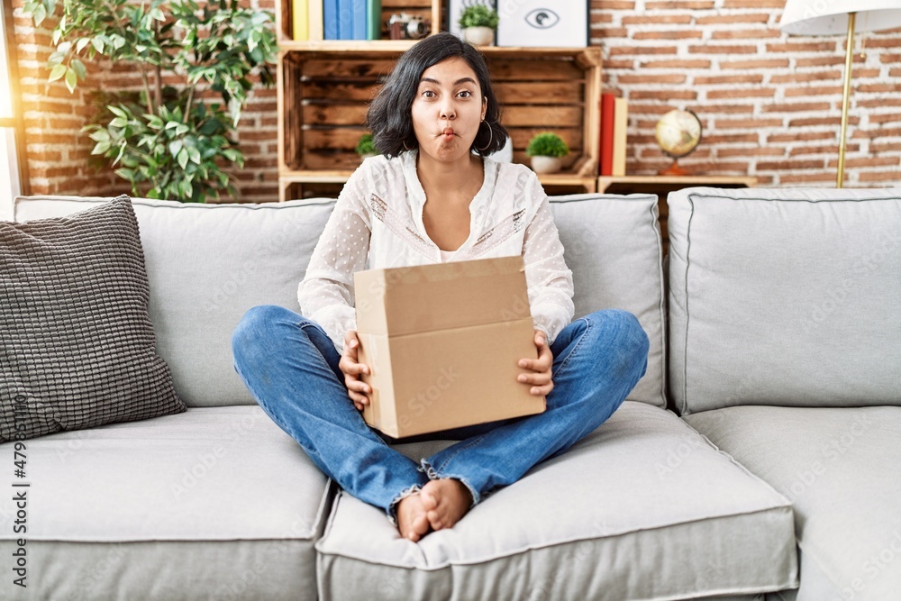 Young hispanic woman sitting on the sofa checking delivery package making fish face with mouth and squinting eyes, crazy and comical. Young hispanic woman sitting on the sofa checking delivery package making fish face with mouth and squinting eyes, crazy and comical.