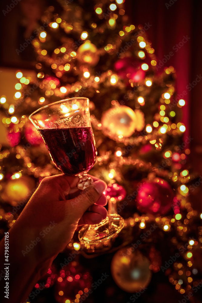 Hand cheering with tilted red wine glass against out of focus christmas tree lights, xmas festive celebration concept.
