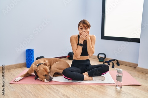 Young beautiful woman sitting on yoga mat shocked covering mouth with hands for mistake. secret concept.