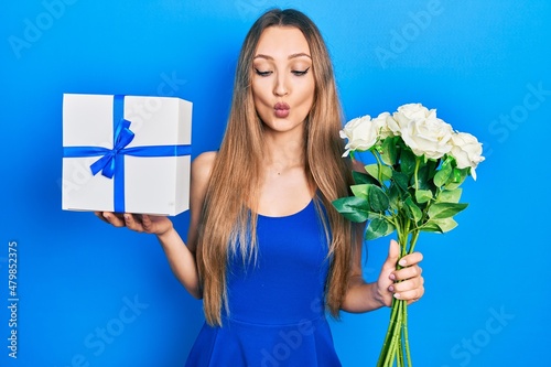 Young blonde girl holding anniversary present and bouquet of flowers making fish face with mouth and squinting eyes, crazy and comical.