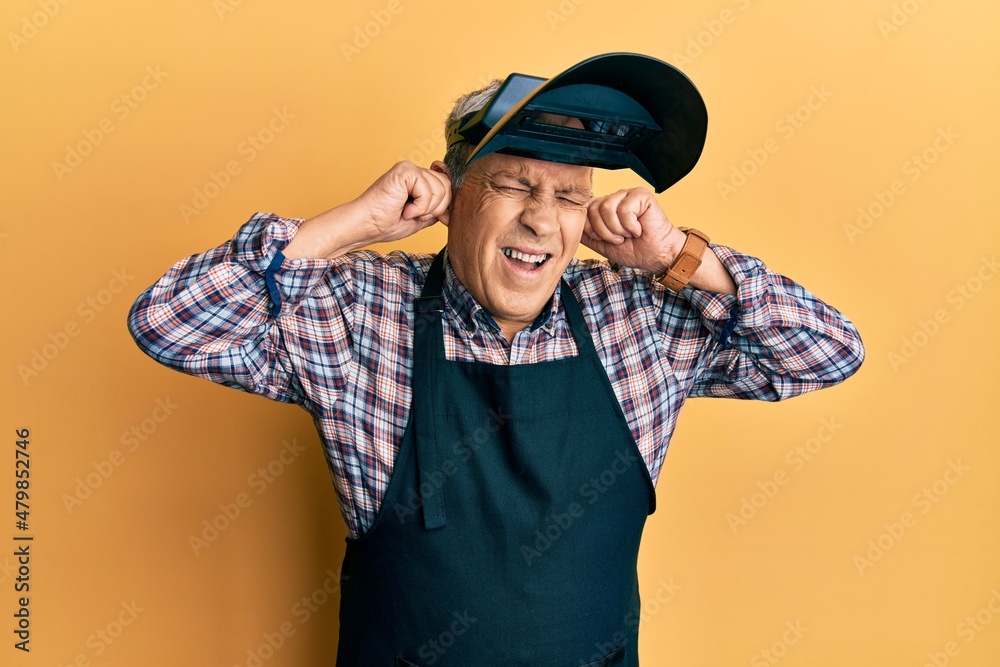 Handsome senior man with grey hair wearing welding protection mask covering ears with fingers with annoyed expression for the noise of loud music. deaf concept.
