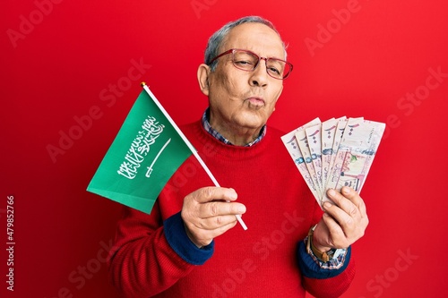 Handsome senior man with grey hair holding saudi arabia flag and riyal banknotes looking at the camera blowing a kiss being lovely and sexy. love expression.