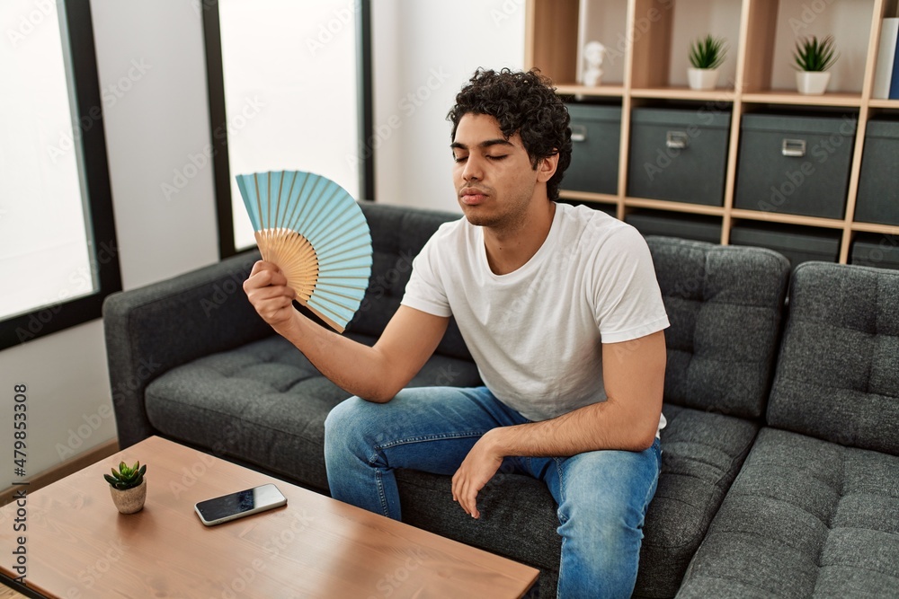 Young hispanic man using hand fan sitting on the sofa at home.