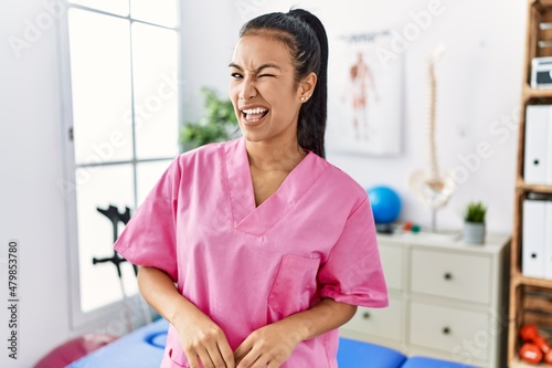 Young hispanic woman working at pain recovery clinic winking looking at the camera with sexy expression, cheerful and happy face.