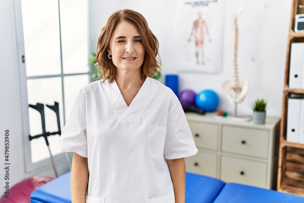 Fototapeta premium Middle age physiotherapist woman working at pain recovery clinic winking looking at the camera with sexy expression, cheerful and happy face.