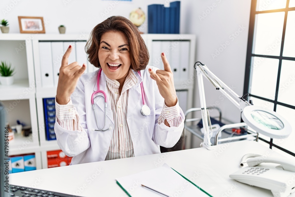 Middle age hispanic woman wearing doctor uniform and stethoscope at the clinic shouting with crazy expression doing rock symbol with hands up. music star. heavy concept. Middle age hispanic woman wearing doctor uniform and stethoscope at the clinic shouting with crazy expression doing rock symbol with hands up. music star. heavy concept.