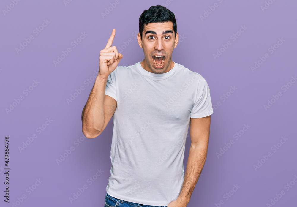 Handsome hispanic man wearing casual white t shirt pointing finger up with successful idea. exited and happy. number one. Handsome hispanic man wearing casual white t shirt pointing finger up with successful idea. exited and happy. number one.