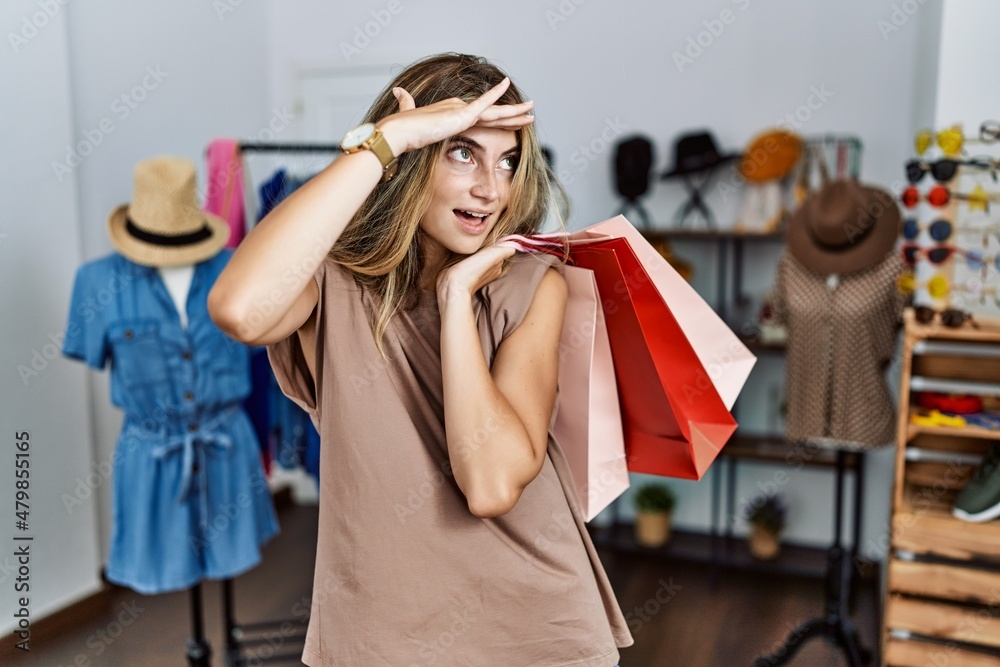 Young blonde woman holding shopping bags at retail shop very happy and smiling looking far away with hand over head. searching concept. Young blonde woman holding shopping bags at retail shop very happy and smiling looking far away with hand over head. searching concept.
