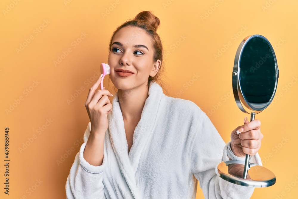 Young blonde woman wearing robe holding makeup remover brush smiling looking to the side and staring away thinking. Young blonde woman wearing robe holding makeup remover brush smiling looking to the side and staring away thinking.