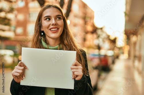Young blonde girl smiling happy holding empty blank banner at the city.