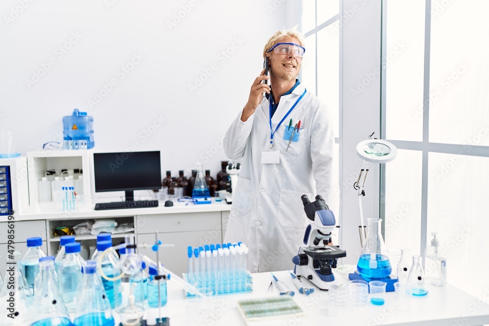Fototapeta premium Young caucasian man wearing scientist uniform talking on the smartphone at laboratory
