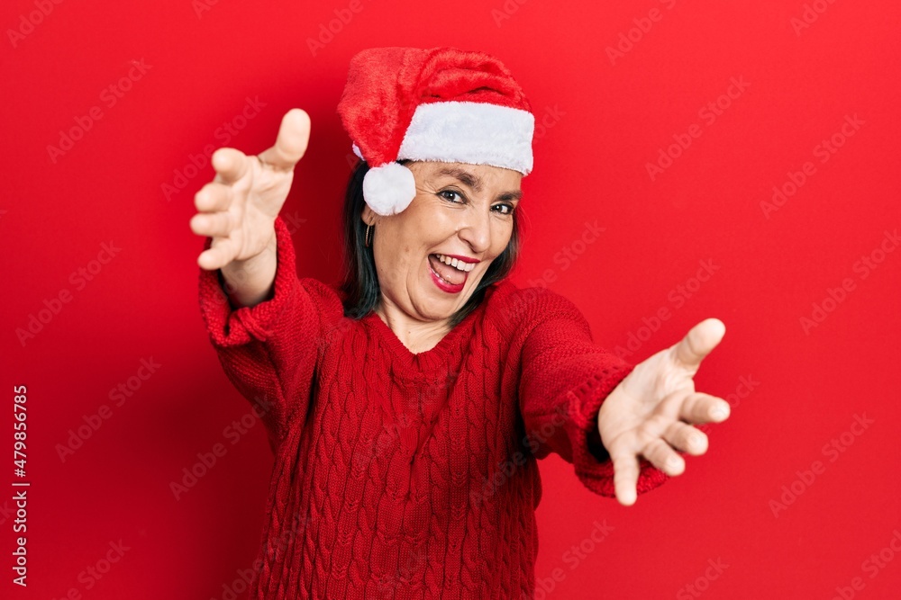 Middle age hispanic woman wearing christmas hat looking at the camera smiling with open arms for hug. cheerful expression embracing happiness. Middle age hispanic woman wearing christmas hat looking at the camera smiling with open arms for hug. cheerful expression embracing happiness.
