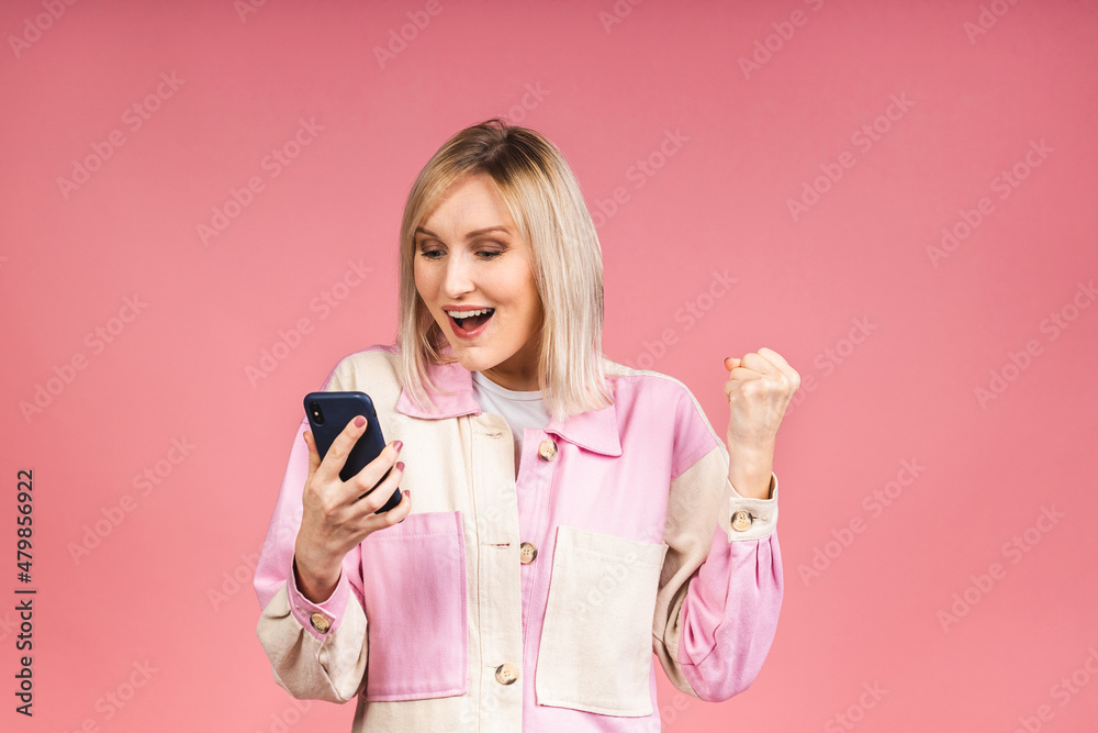 Happy winner. Blonde young female with terrible expression in smartphone, reads shocking news on web page, isolated on pink background. Shocked amazed woman using mobile phone. Happy winner. Blonde young female with terrible expression in smartphone, reads shocking news on web page, isolated on pink background. Shocked amazed woman using mobile phone.