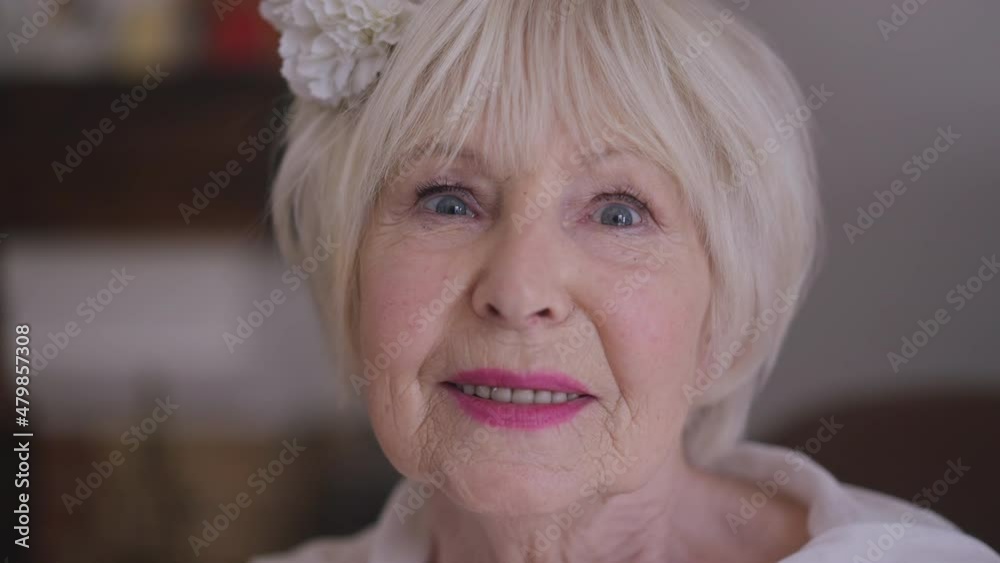Headshot of gorgeous Caucasian senior woman in wedding dress with flower in hair smiling looking at camera. Close-up confident happy bride posing in slow motion indoors at home. Beauty and femininity Headshot of gorgeous Caucasian senior woman in wedding dress with flower in hair smiling looking at camera. Close-up confident happy bride posing in slow motion indoors at home. Beauty and femininity