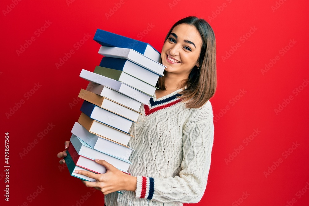 Young brunette student girl holding a pile of books smiling with a happy and cool smile on face. showing teeth. Young brunette student girl holding a pile of books smiling with a happy and cool smile on face. showing teeth.