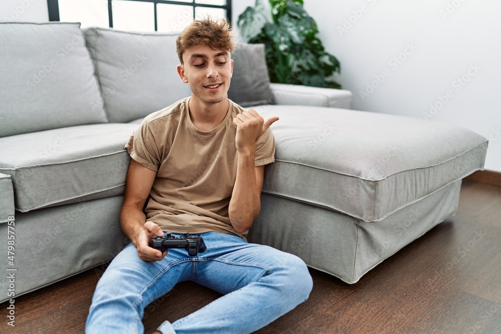 Young caucasian man playing video game holding controller at home pointing thumb up to the side smiling happy with open mouth