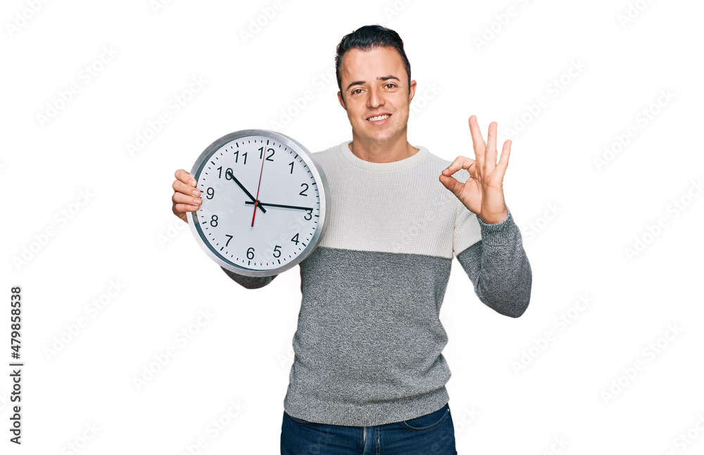 Handsome young man holding big clock doing ok sign with fingers, smiling friendly gesturing excellent symbol Handsome young man holding big clock doing ok sign with fingers, smiling friendly gesturing excellent symbol