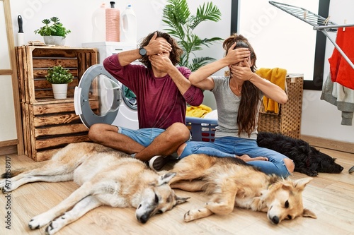 Photography Young hispanic couple doing laundry with dogs covering eyes and mouth with hands, surprised and shocked
