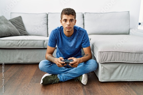Fotografie Young handsome hispanic man playing video game sitting by the sofa thinking atti