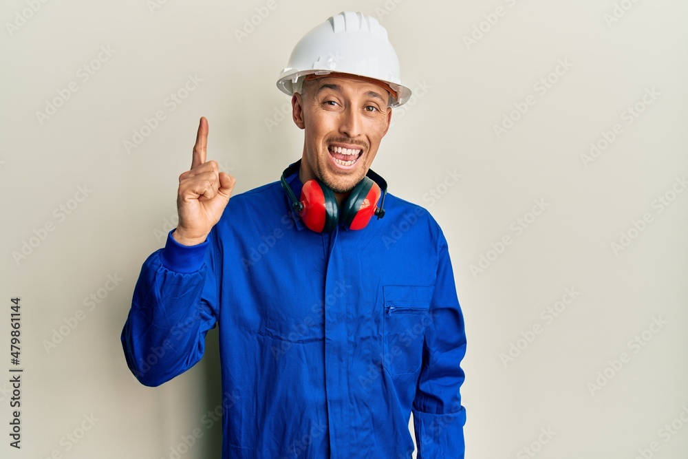 Bald man with beard wearing builder jumpsuit uniform and hardhat pointing finger up with successful idea. exited and happy. number one.