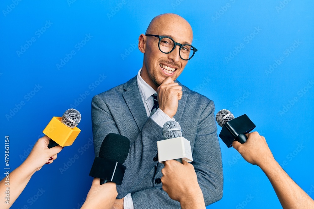 Bald man with beard being interviewed by reporters holding microphones smiling looking confident at the camera with crossed arms and hand on chin. thinking positive. Bald man with beard being interviewed by reporters holding microphones smiling looking confident at the camera with crossed arms and hand on chin. thinking positive.