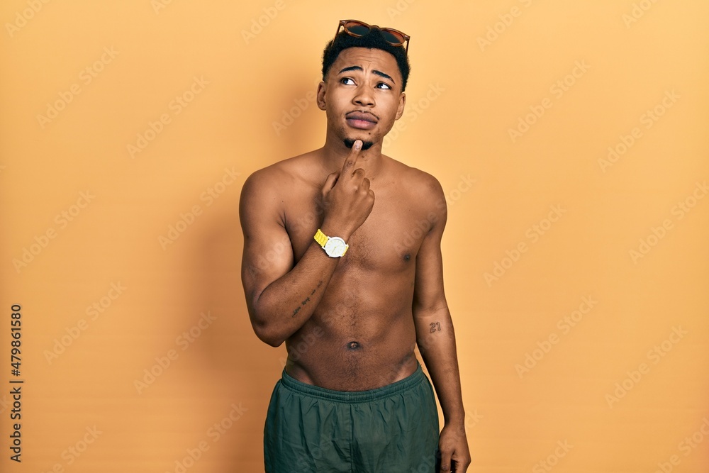 Young african american man wearing swimwear and swimmer glasses thinking concentrated about doubt with finger on chin and looking up wondering