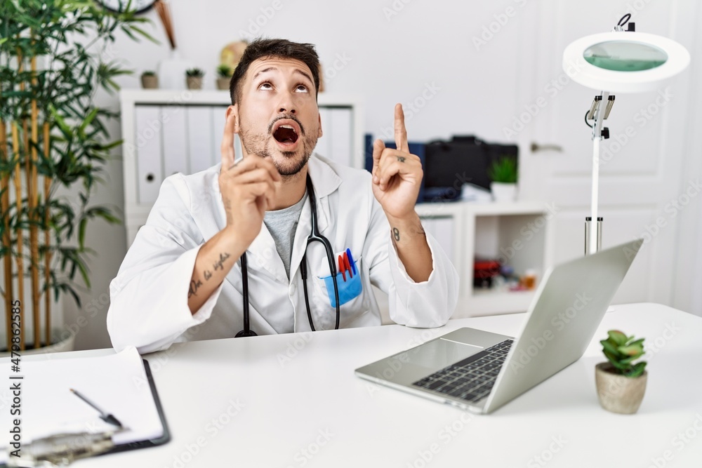 Young doctor working at the clinic using computer laptop amazed and surprised looking up and pointing with fingers and raised arms.