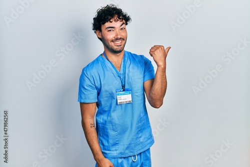 Young hispanic man wearing blue male nurse uniform smiling with happy face looking and pointing to the side with thumb up.