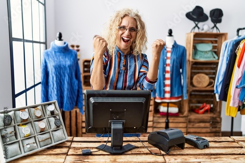 Middle age blonde woman working as manager at retail boutique very happy and excited doing winner gesture with arms raised, smiling and screaming for success. celebration concept.
