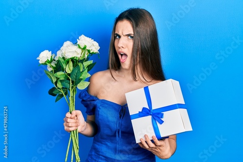 Young brunette teenager holding anniversary present and bouquet of flowers in shock face, looking skeptical and sarcastic, surprised with open mouth