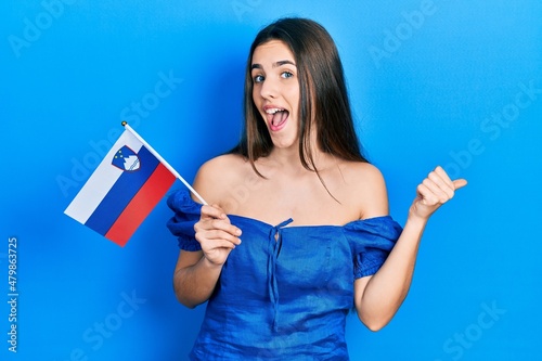 Young brunette teenager holding slovenia flag pointing thumb up to the side smiling happy with open mouth