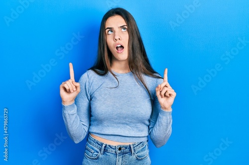 Young brunette teenager wearing casual sweater amazed and surprised looking up and pointing with fingers and raised arms.