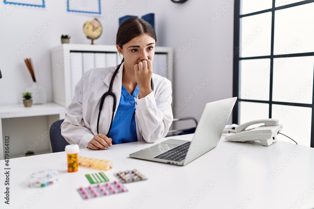 Young hispanic doctor woman wearing doctor uniform working at the clinic looking stressed and nervous with hands on mouth biting nails. anxiety problem.